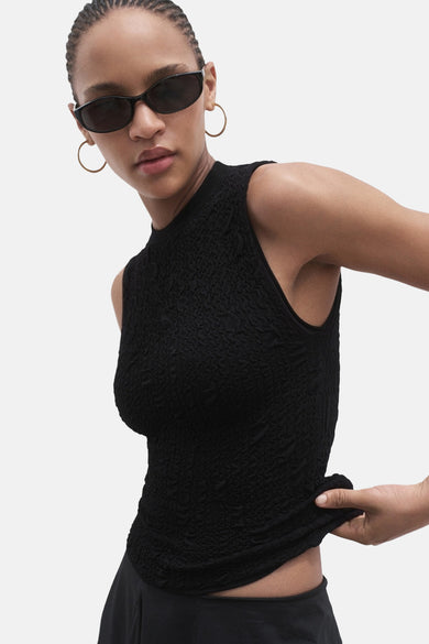 Blouse sans manches noire de MANGO, avec une texture froissée et portée avec un chapeau de soleil et des boucles d'oreilles en or.