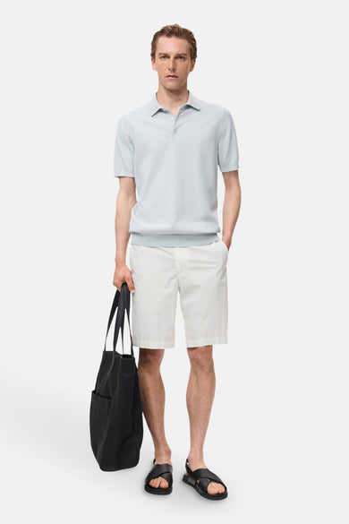 Polo bleu clair à manches courtes de MANGO, porté avec un short blanc, une sacoche noire et des sandales.