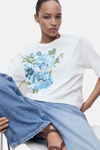 T-shirt blanc à manches courtes de MANGO, imprimé de fleurs bleues, porté avec un pantalon de jeans large.
