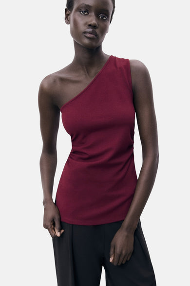 T-shirt sans manches bordeaux de MANGO, avec une ligne d'épaules asymétrique et porté avec un pantalon noir.