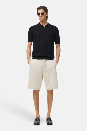 Ecru jeansshort van MANGO, gecombineerd met een zwart poloshirt, zonnebril en leren loafers.
