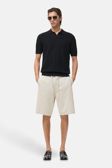 Jean écru court de MANGO, associé à un t-shirt polo noir, des lunettes de soleil et des loafers en cuir.
