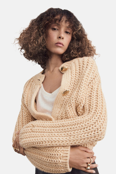 Beige gilet van MANGO, grof gebreid met gouden knopen en gedragen over een wit shirt.