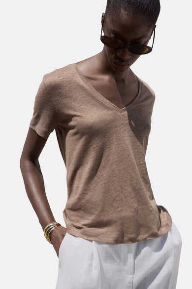 T-shirt marron à manches courtes de MANGO, avec un col V, associé à une pantalon blanc et des bracelets dorés.