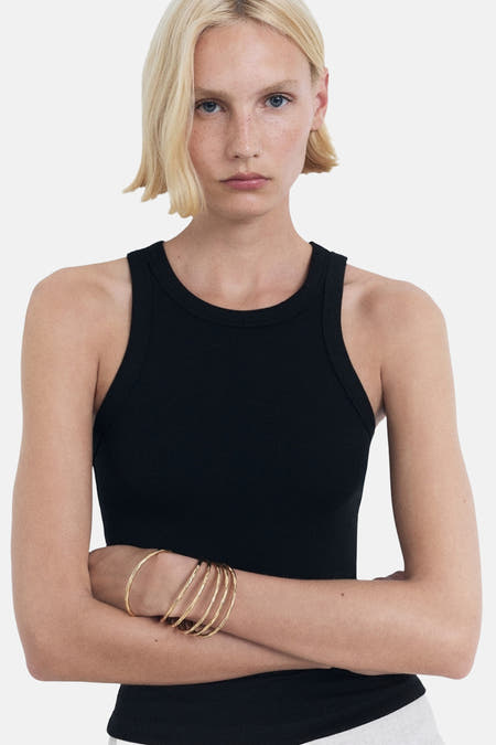 Zwarte singlet van MANGO, met gekruiste armen met gouden armbanden, tegen een witte achtergrond.