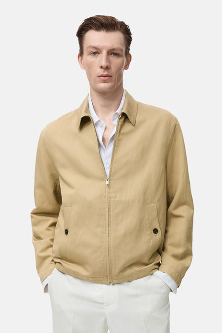 Beige korte jas van MANGO, met rits en steekzakken, gedragen over een wit overhemd en witte broek.
