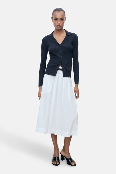Cardigan bleu de MANGO, porté avec une jupe blanche et des escarpins noirs.