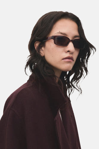 Bordeaux long manteau de MANGO, porté avec un chapeau de soleil, contre un fond neutre.