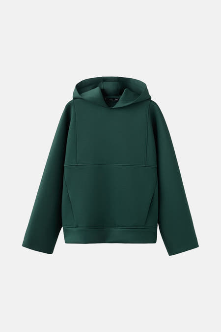 Pull-over vert de MANGO, porté sous un manteau en laine à carreaux et une chemise bleu clair.