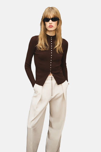 Gilet marron de MANGO, avec fermeture à boutons, associé à un pantalon crème avec une taille haute et une fermeture éclair visible.