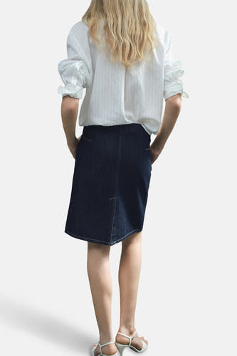 Donkerblauwe denim rok van MANGO, met een asymmetrische zoom en achterzakken, gedragen met een gestreept overhemd en witte sandalen.
