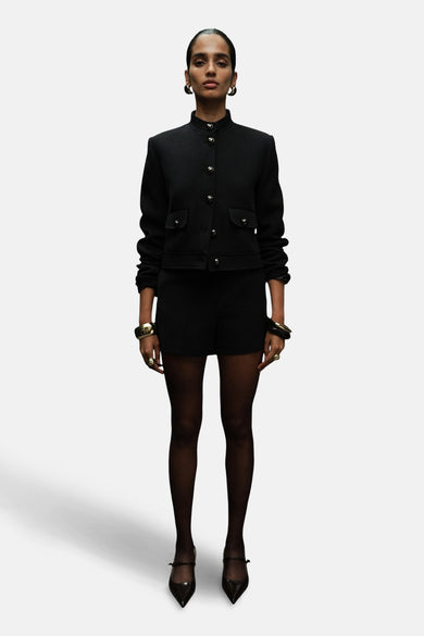 Zwarte blazer van MANGO, met gouden knopen en opgestikte zakken, gedragen met zwarte shorts en panty's.
