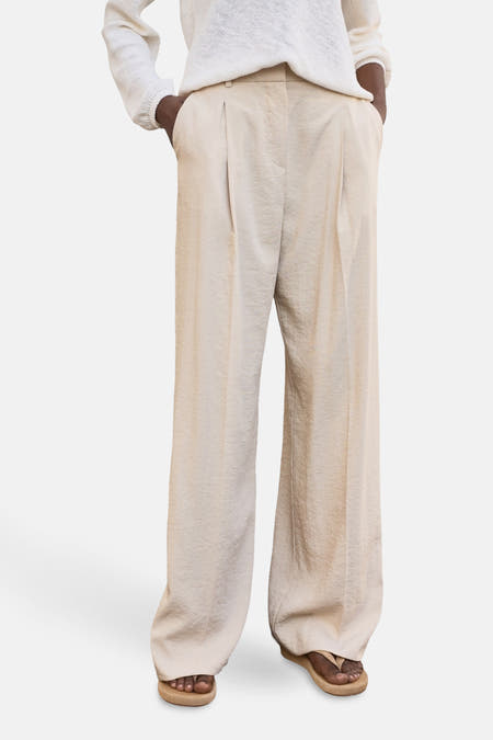 Beige broek van MANGO, met wijde pijpen en gedrapeerde textuur, gedragen met sandalen.
