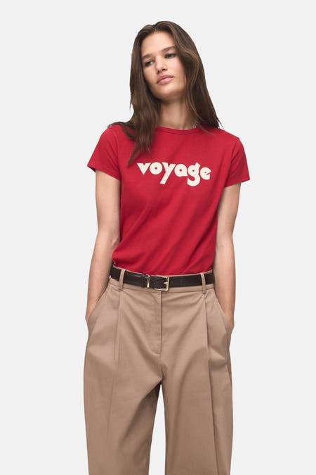 Rood T-shirt met korte mouwen van MANGO, met de tekst 'voyage' in witte letters, gedragen met een bruine broek.
