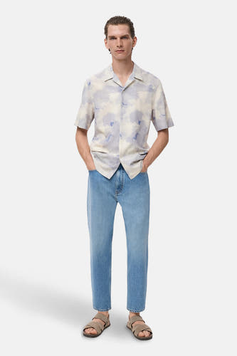 Multikleurig overhemd met korte mouwen van MANGO, met een abstract wolkenpatroon, gecombineerd met lichtblauwe jeans en sandalen.
