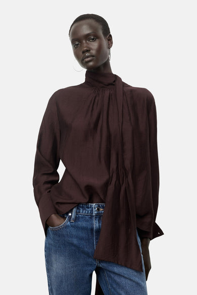 Bruine blouse met lange mouwen van MANGO, met hoge hals en gedrapeerde details gecombineerd met blauwe jeans.
