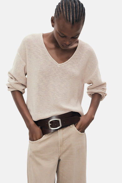 Gilet V-hals beige de MANGO, avec des manches longues et une coupe décontractée, associé à un ceinture brune et une pantalon beige.