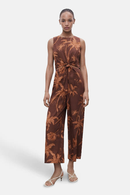 Bruine jumpsuit van MANGO met palmboomprint en knoopdetail aan de voorkant, gedragen met witte sandalen.