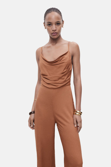 Bruine jumpsuit van MANGO, met een gedrapeerde halslijn en gouden armbanden.
