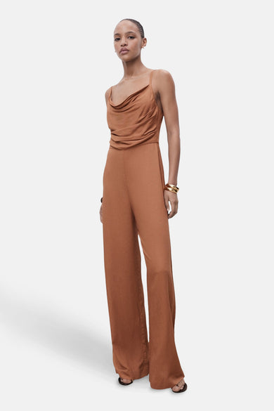 Bruine jumpsuit van MANGO met een gedrapeerde halslijn en wijde pijpen, gecombineerd met gouden armbanden.
