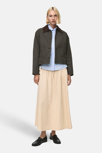Khaki korte jas van MANGO, met bruine kraag, gedragen met een lichtblauw overhemd en een beige rok.
