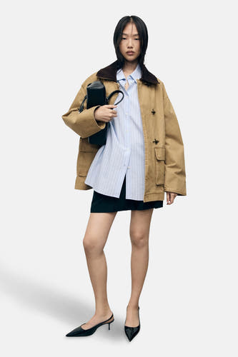 Beige korte jas van MANGO, met bruine kraag, gedragen over een blauw gestreept overhemd en een zwarte rok.