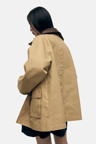 Beige korte jas van MANGO, met bruine kraag en zak op de voorkant, op een model van achteren gezien.

