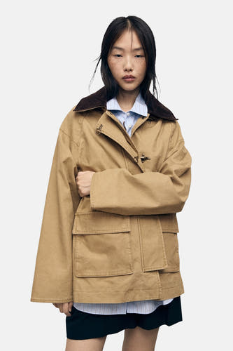 Beige korte jas van MANGO, met bruine kraag en zakken aan de voorkant, gedragen over een gestreept overhemd.
