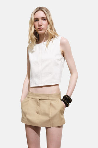 Ecru mouwloze blouse van MANGO, gedragen met een kaki minirok en zwarte armbanden met gouden details.
