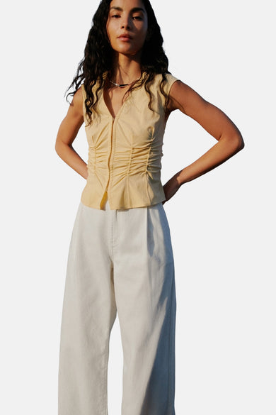 Gele mouwloze blouse van MANGO, met plooien aan de voorkant, gecombineerd met een witte broek.
