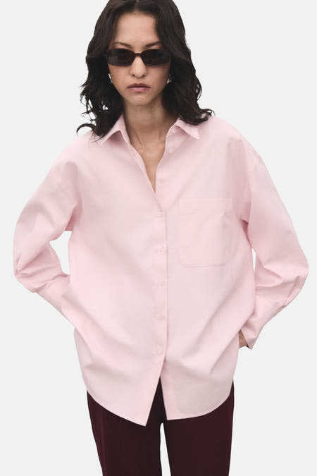 Chemise rose à manches longues de MANGO, avec une poche poitrine et portéeée un pantalon sombre et un chapeau de soleil.