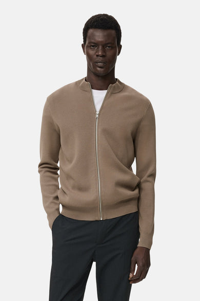 Bruin gilet van MANGO, met lange mouwen en een zilverkleurige ritssluiting, gedragen over een wit t-shirt en een zwarte pantalon.
