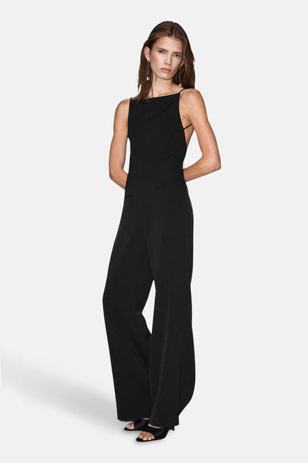Zwarte jumpsuit van MANGO, met brede pijpen en open rugdetail, gedragen met zilveren oorbellen en zwarte hakken.

