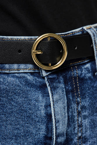 Zwarte riem van PIECES, met een opvallende gouden ronde gesp, gedragen over een blauwe jeans.
