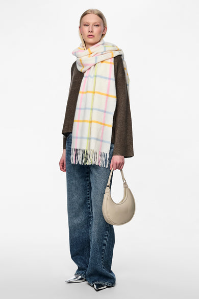 Ecru wintersjaal van PIECES® met pastelkleurige ruit en franjes, gedragen met een bruine trui, jeans en zilverkleurige schoenen.
