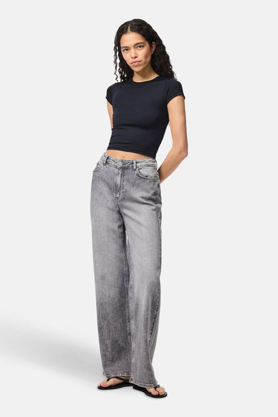 Jean gris large de PIECES®, porté avec un haut noir et des sandales, le modèle est droit les les mains derrière le dos.