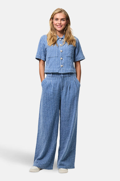 Jean bleu de PIECES, avec des jambes larges et des plis, porté avec une blouse coordonnée.