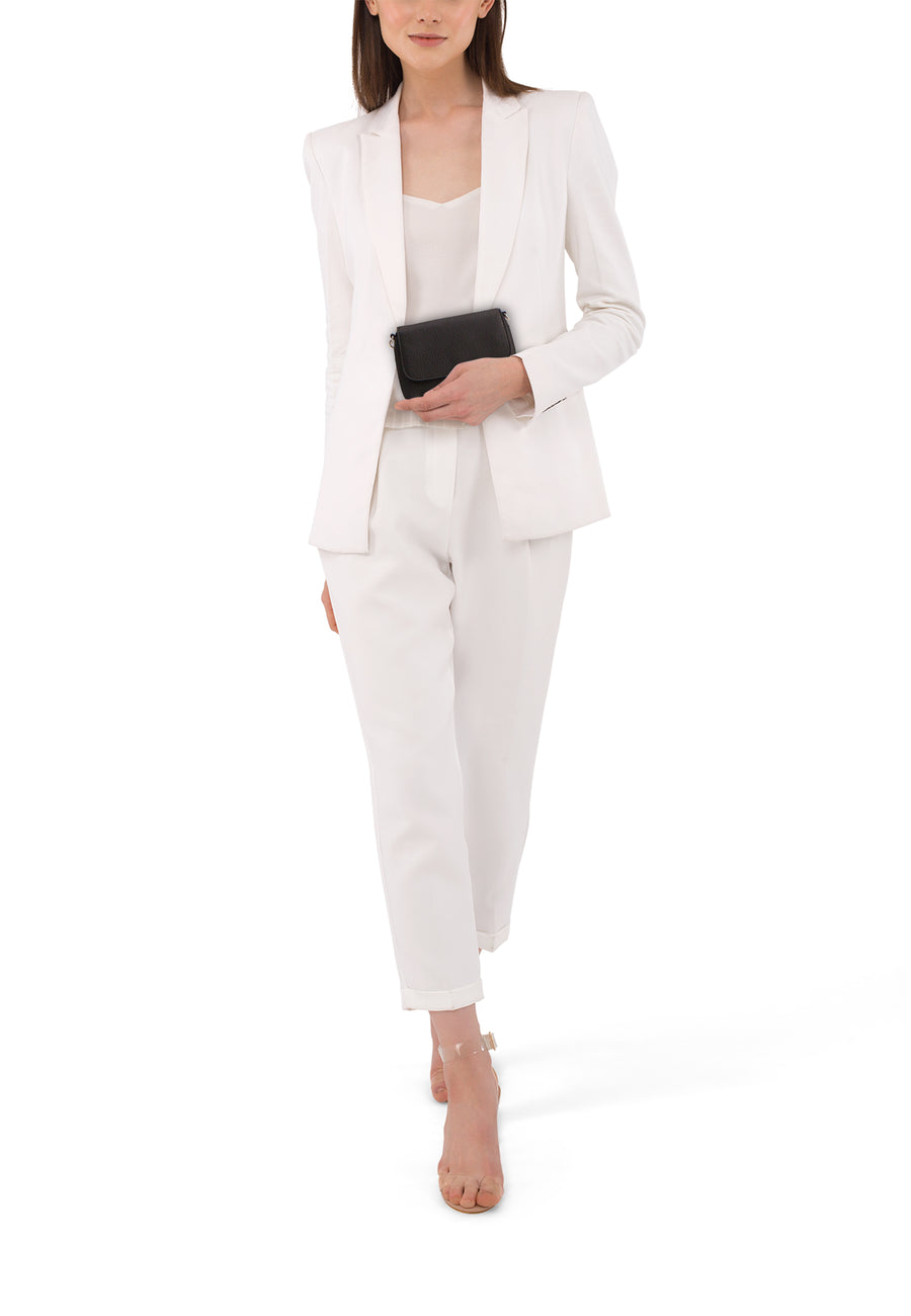 Vrouw in witte broek en blazer met zwarte handtas en transparante hakken, staand op witte achtergrond.