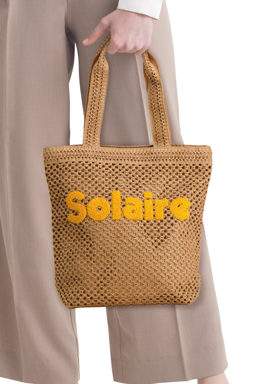 Sac à main tissé marron de PIECES avec texte jaune 'Solaire', porté par une personne en pantalon beige.