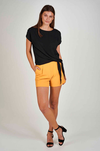 Gele short van Morgan De Toi, gecombineerd met een zwarte blouse met knoopdetail en zwarte hakken.
