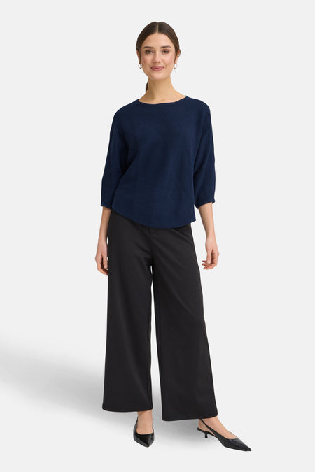 Vrouw met donkerblauwe trui, zwarte broek en zwarte pumps, staand voor een witte achtergrond.