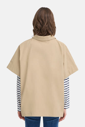 Manteau beige de France, avec des manches courtes, porté sur une chemise rayée et un jean bleu.