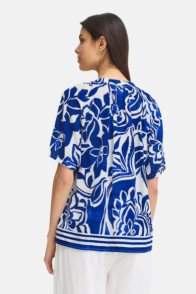 Blouse bleue Fransa® avec imprimé de fleurs et manches courtes, vue de l'arrière, associée à un pantalon blanc.