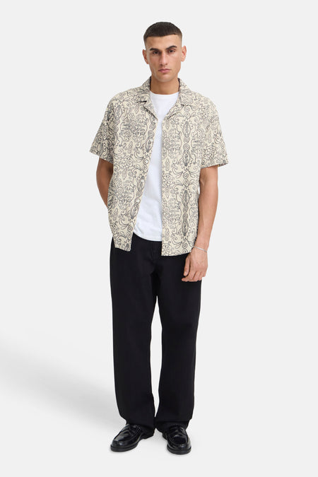 Ecru overhemd met korte mouwen van SOLID met een zwart abstract bloemenpatroon, gedragen over een wit T-shirt, zwarte broek en loafers.