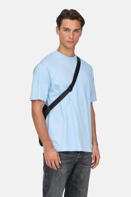 T-shirt bleu à manches courtes de ONLY & SONS®, porté avec une sacoche noire et des jeans gris.