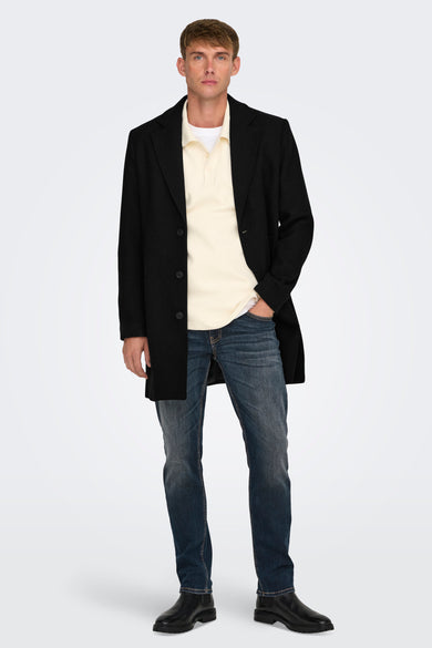 Lange zwarte jas van ONLY & SONS, gedragen over een crèmekleurige polo en donkerblauwe jeans.