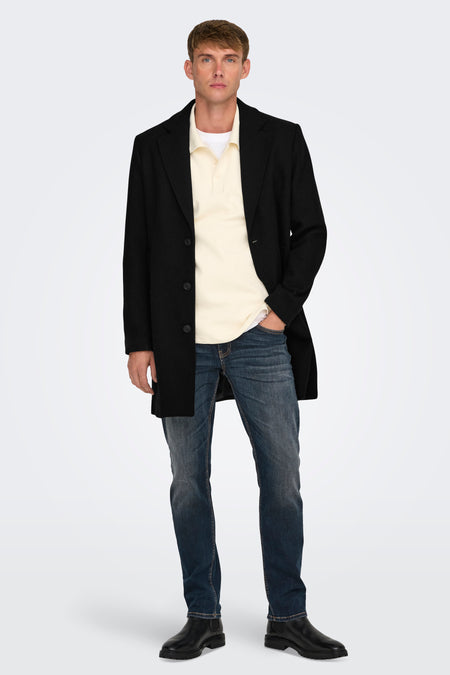 Lange zwarte jas van ONLY & SONS, gedragen over een crèmekleurige polo en donkerblauwe jeans.