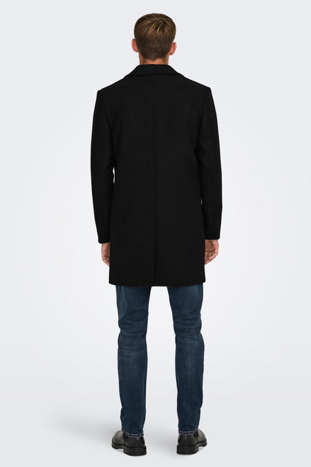 Zwarte lange jas van ONLY & SONS, met een effen patroon, gedragen over een donkere spijkerbroek en zwarte schoenen, achterkant.
