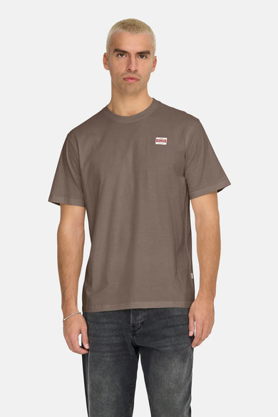 Bruin T-shirt met korte mouwen van ONLY & SONS, met een klein merklogo op de borst, gecombineerd met donkere jeans en een zilveren armband.
