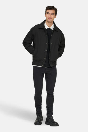 Zwarte jas van ONLY & SONS, met lange mouwen, knoopsluiting en zakken, gedragen over een wit overhemd, een zwarte trui en een zwarte jeans.